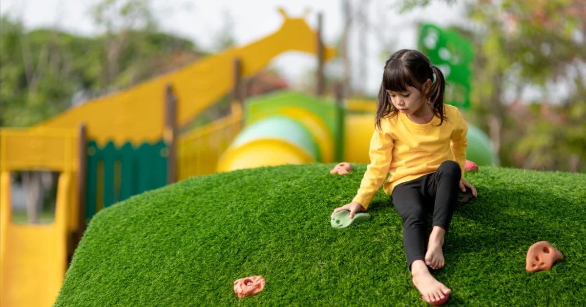 Young girl climbing a green synthetic turf hill in a playground with colorful climbing holds